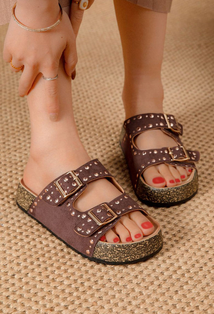 Brown sandals with glittery soles worn by a person on a textured surface