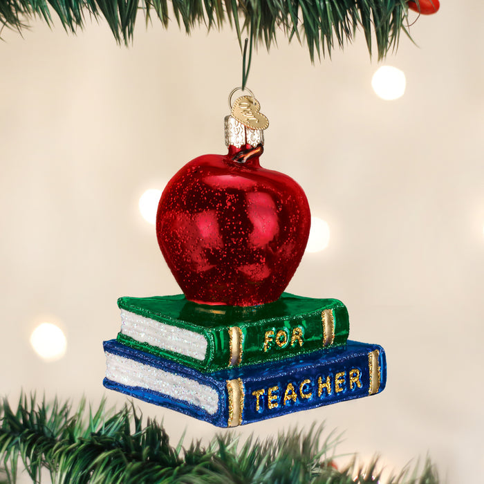A shiny red apple ornament sits atop two stacked books labeled FOR and TEACHER, hanging from a green Christmas tree branch with blurred lights in the background.