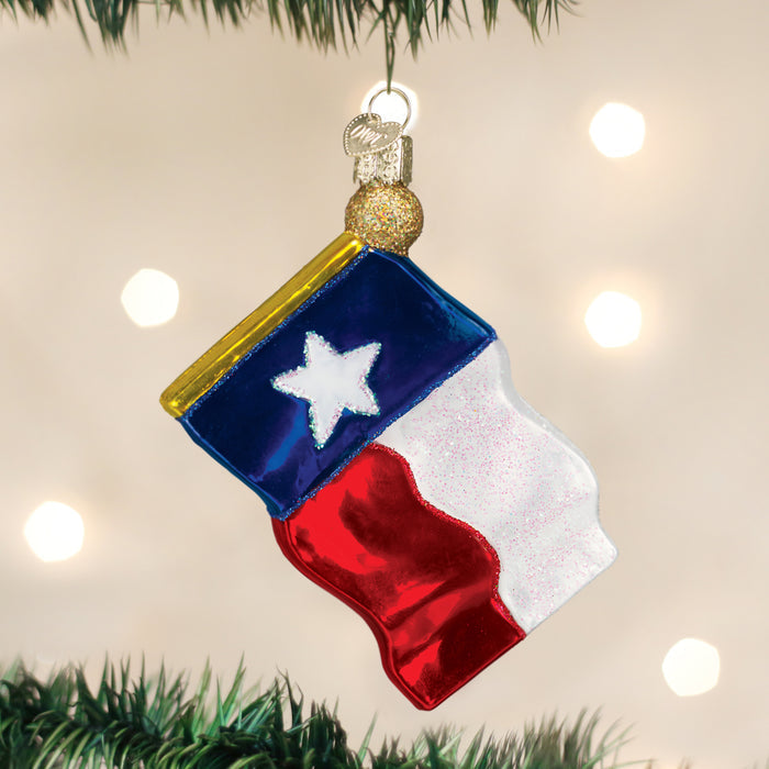 A Christmas tree ornament shaped and colored like the Texas state flag hangs from a tree branch, with a blurred background of white lights.
