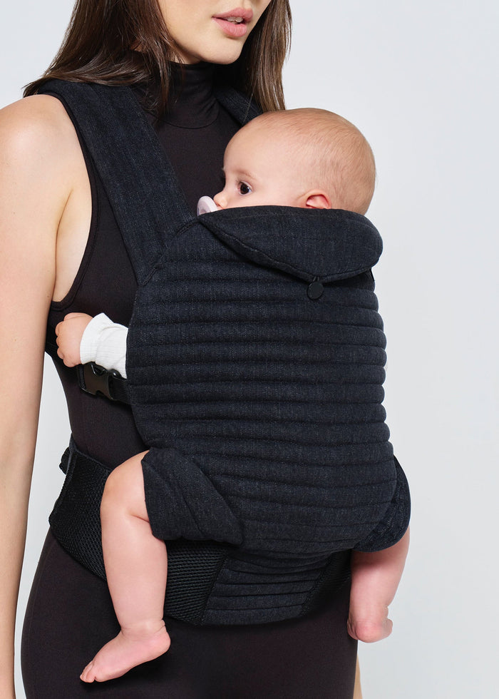 Woman holding a baby in a black  denim baby carrier by Bumpsuit against a white background