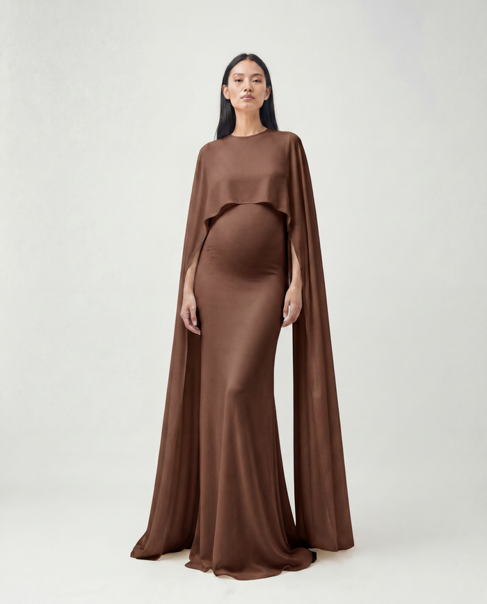 A pregnant model wearing a Bumpsuit brown maternity gown with a draped cape overlay, standing against a light gray studio background.