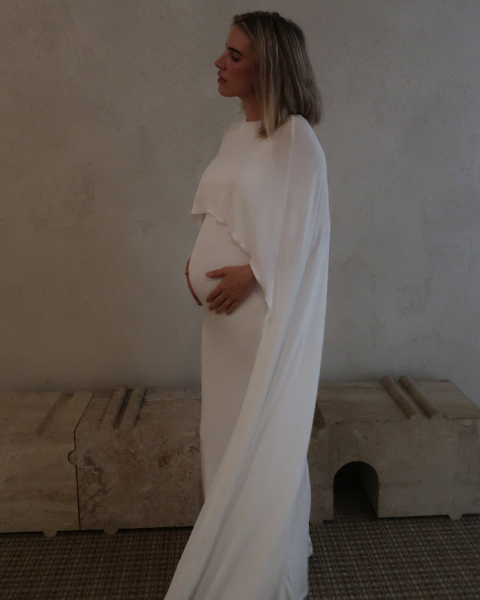 Side view of a pregnant woman wearing a maxi cape style dress in Ivory  against a beige background