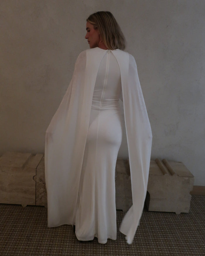 Back view of a woman wearing Bumpsuit maxi gown with a long sheer cape, standing in a neutral indoor setting.