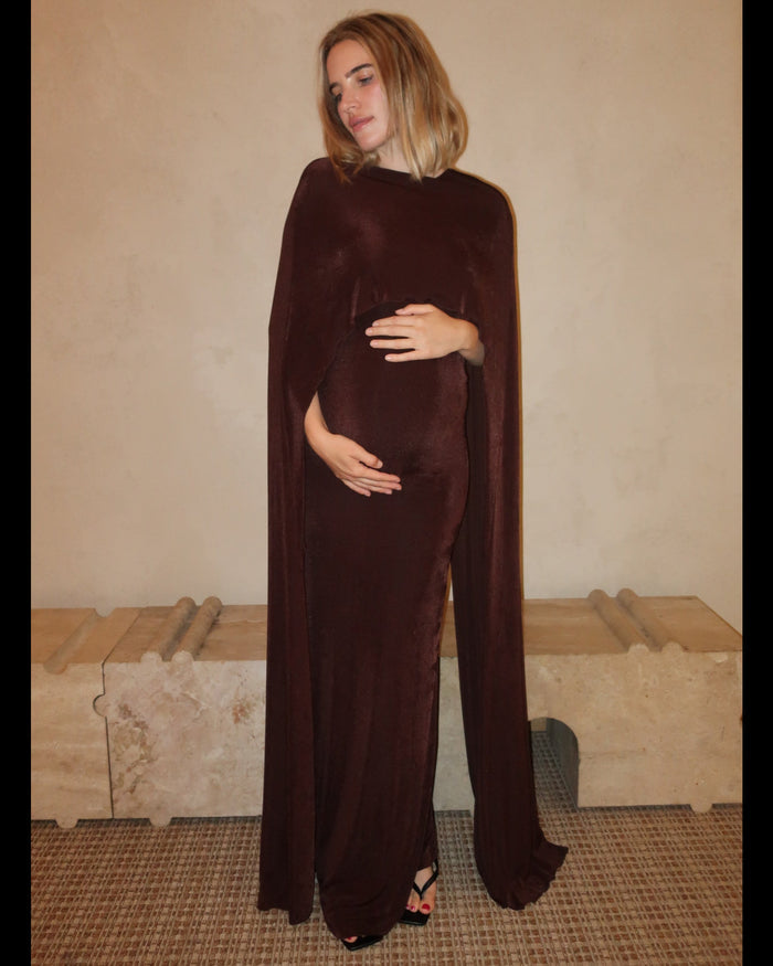 A pregnant woman wearing the Aubrey Bumpsuit maxi dress with a floor length cape detail in chocolate brown against a neutral setting