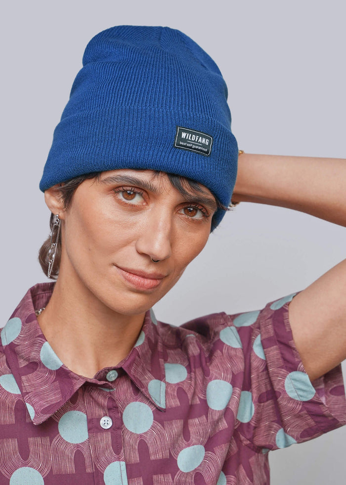 A person wearing a blue ribbed knit beanie with a small black rectangular brand patch that reads 'WILDFANG' and a mauve short-sleeve button-up shirt with a light blue and mauve geometric pattern, against a plain light gray studio background.
