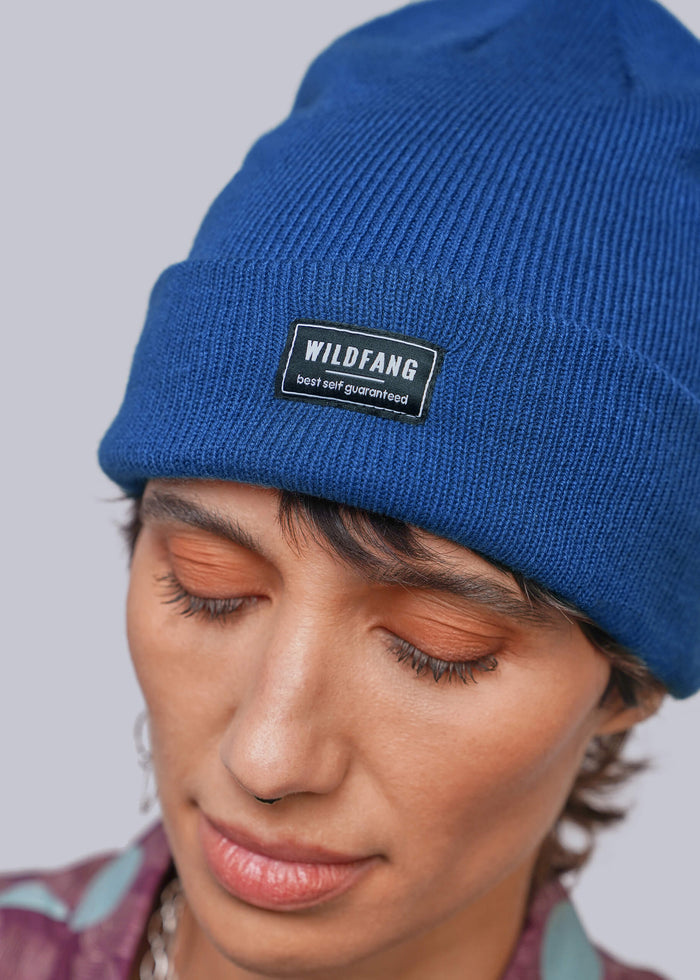 A close-up of a person wearing a royal blue ribbed knit beanie with a black rectangular patch that reads 'WILDFANG best self guaranteed.' The person is shown from the shoulders up against a light gray studio background.