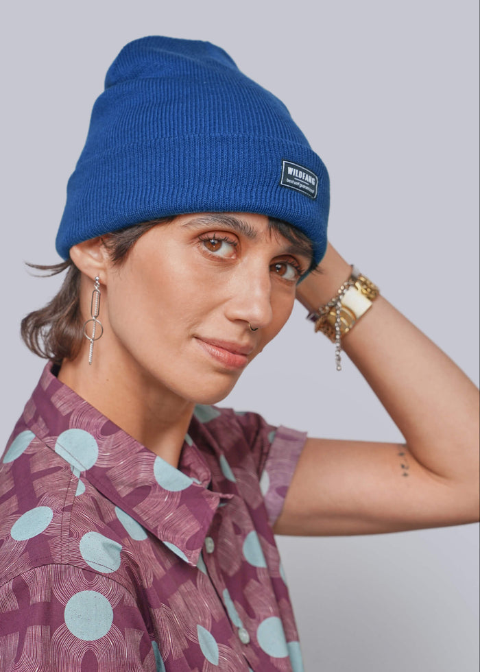 A person wearing a bright blue ribbed knit beanie with a small black brand patch on the front, paired with a purple short-sleeve button-up shirt featuring a light blue geometric pattern. She is posed against a solid light gray background.