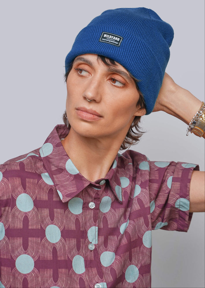 A person wearing a blue ribbed beanie with a small black rectangular logo patch that reads 'WILDFANG' and a patterned short-sleeve button-up shirt, posed against a light gray studio background.