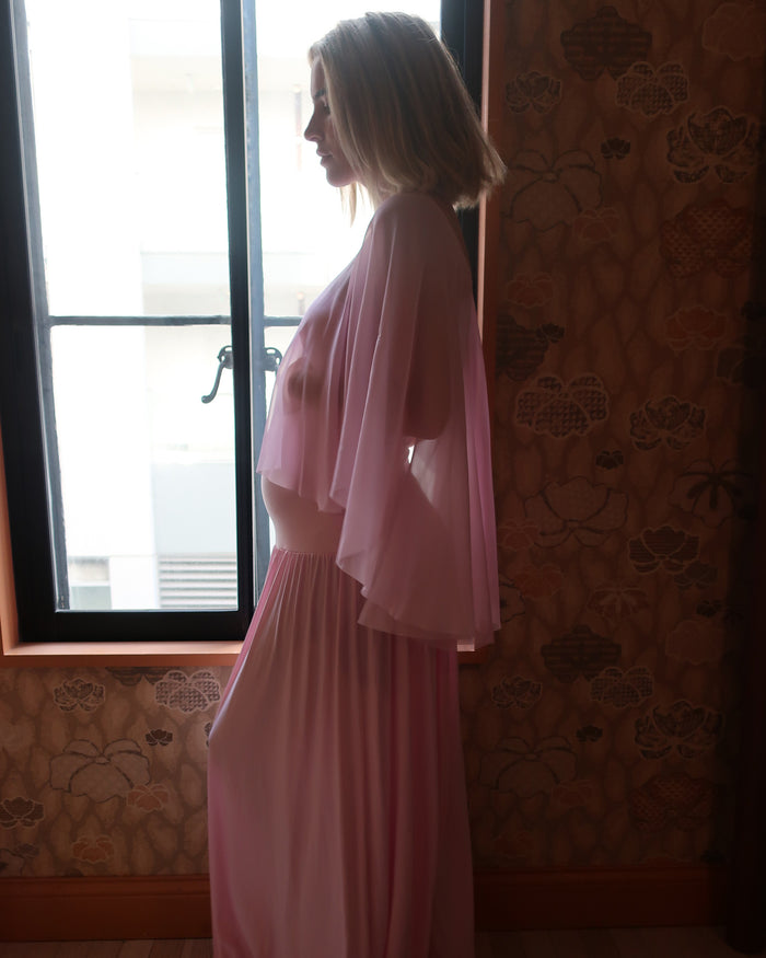A pregnant woman in profile wearing a long pink dress by Bumpsuit with a sheer chiffon cape, standing indoors near a window with patterned wallpaper.