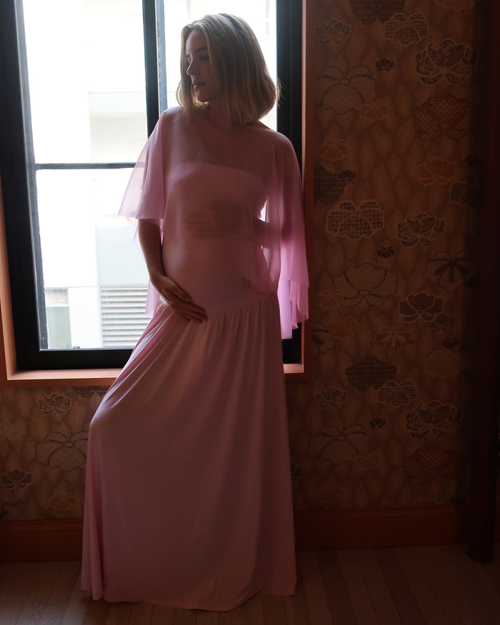 A pregnant woman wearing a Bumpsuit long, flowing light pink gown with a chiffon cape  stands in front of a window, with patterned wallpaper and wood flooring visible in the room.