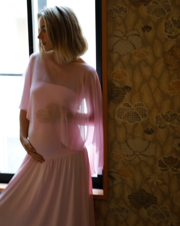 A pregnant woman with blonde hair stands by a window wearing a bumpsuit pink dress with a chiffon cape, infront of a window in a room with patterned wallpaper visible on the right.