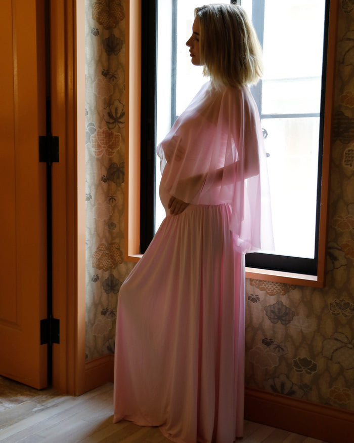 A pregnant woman in a Bumpsuit pink sheer maxi dress with a chiffon cape in profile by a window in a room with floral wallpaper and wood trim.