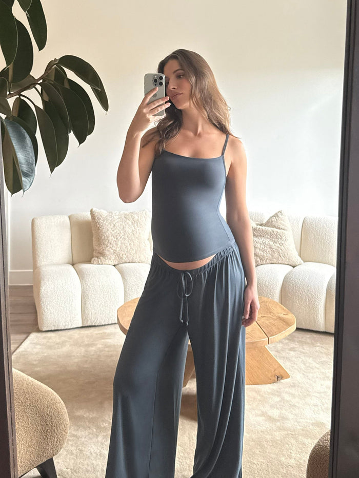 Pregnant woman taking a mirror selfie wearing a Bumpsuit cloud cement blue tank top and pants in a living room.