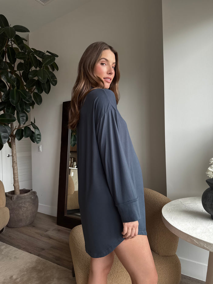 Pregnant woman wearing a Bumpsuit Cloud cement blue pyjama dress standing in a room with a plant and table.