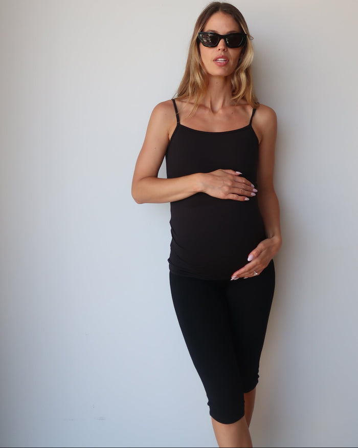 Pregnant woman wearing a black Bumpsuit camisole tank top and capri leggings against a white wall