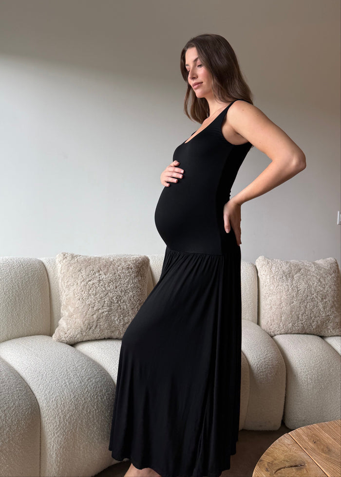 Pregnant woman in a black cotton dress standing in a living room with beige sofa and cushions.