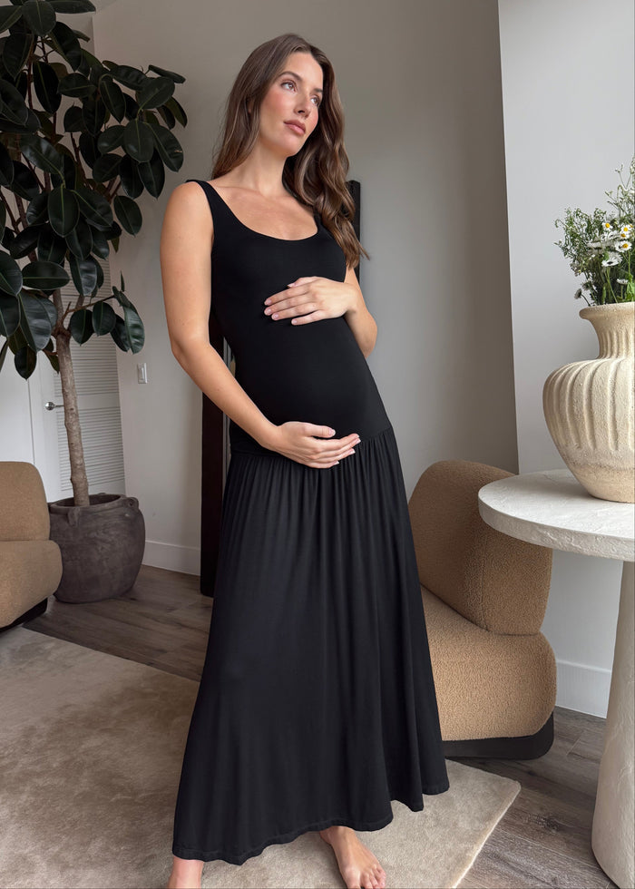 Pregnant woman in a black cotton tank dress by Bumpsuit standing in a modern living room.
