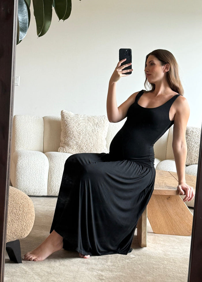 Pregnant woman in a black dress taking a mirror selfie in a living room.