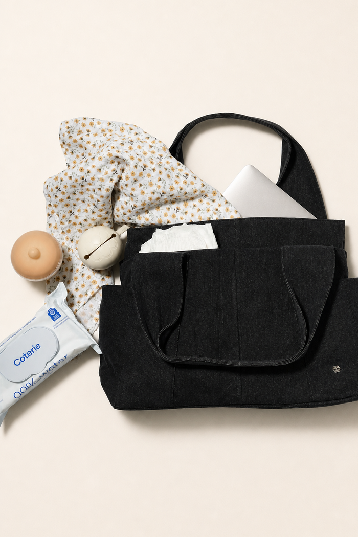 A black denim fabric diaper bag shown in a styled flat-lay with a laptop, patterned scarf, milk bottle, and wipes packet around it on a warm off-white background.
