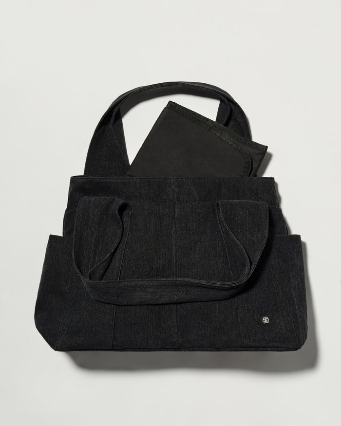 A black denim fabric diaper bag with dual top handles and a matching black fabric change mat insert or pouch partially tucked inside, photographed on a light grey background.