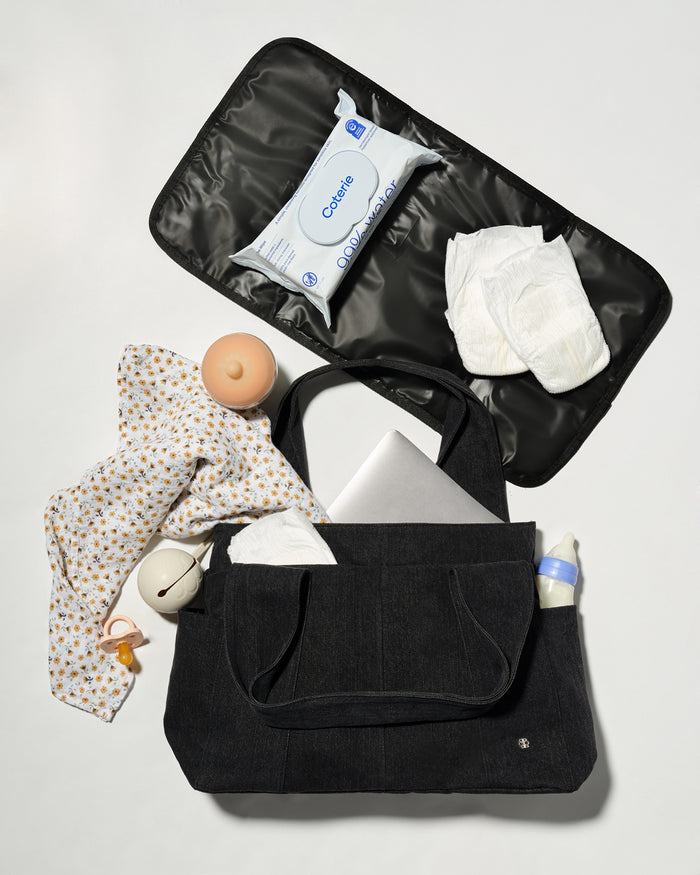 A black diaper bag shown open in a flat-lay arrangement, filled with baby essentials including a patterned baby shirt, wipes, diapers, a pacifier, and a bottle, on a light grey background.