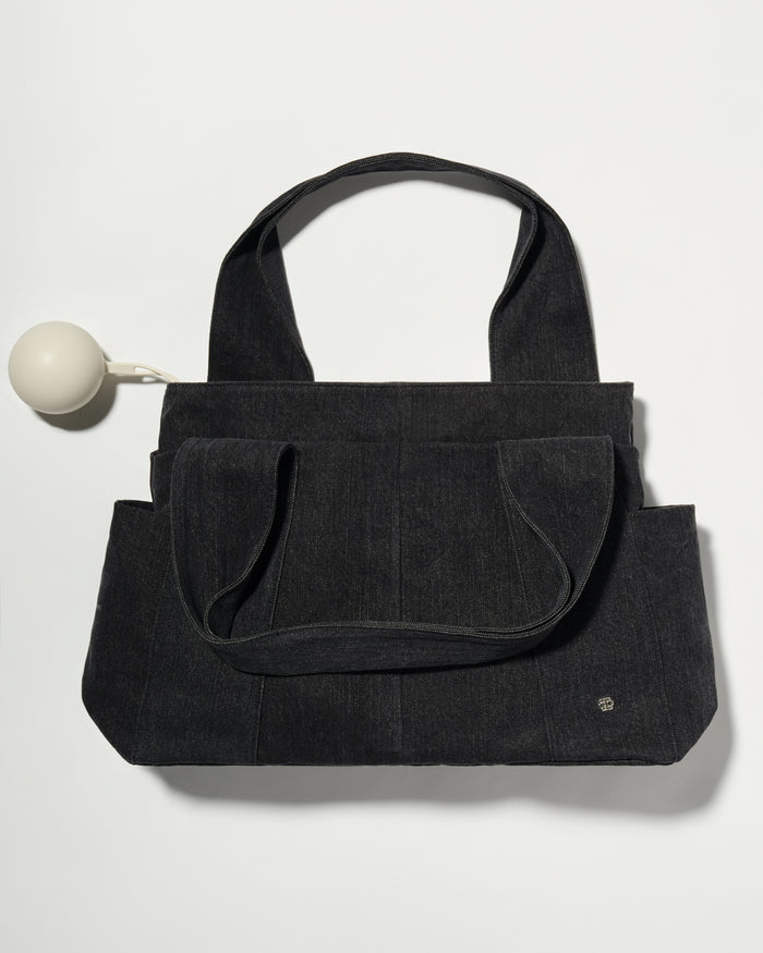 A black denim fabric diaper bag with dual top handles and front slip pockets, shown on a plain light background with a small pacifier holder on the left.