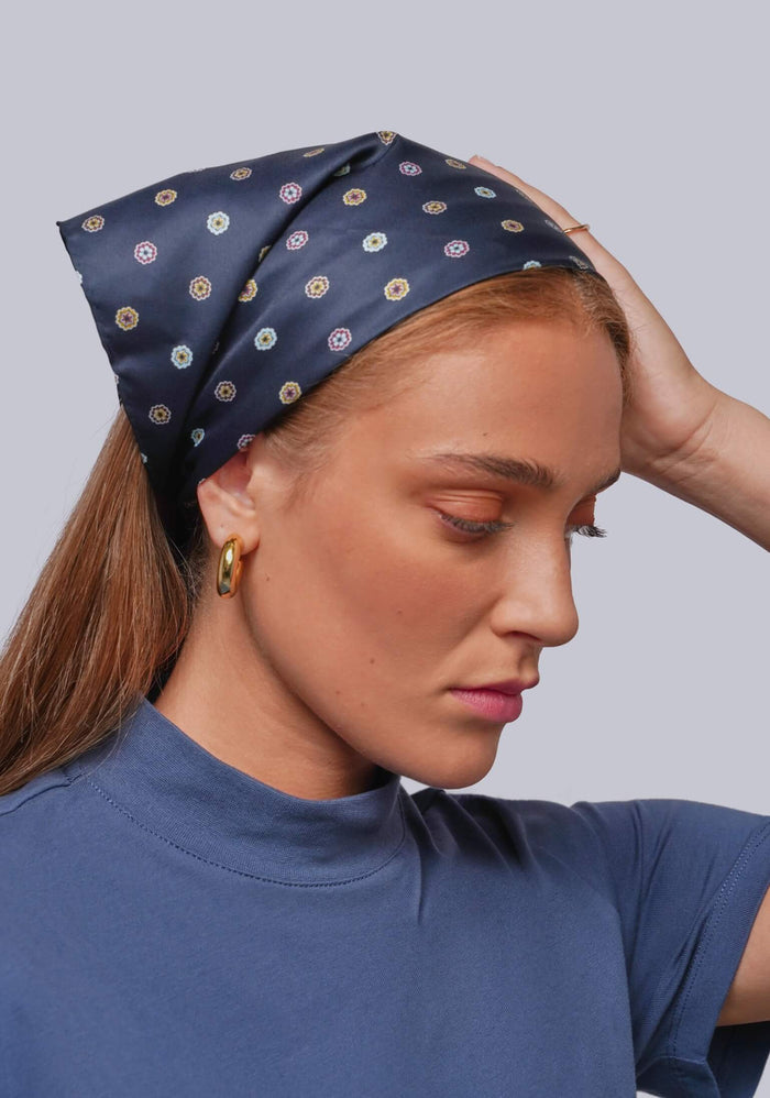 A woman wearing a navy blue silk-like headscarf with a repeating circular pattern in beige, white, and muted red, paired with a blue t-shirt against a solid gray background.