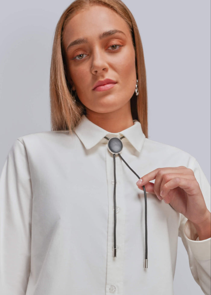 A woman wearing a white long-sleeve button-up shirt with a black bolo tie featuring a round pendnat, posed against a light gray studio background.