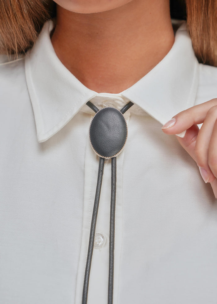 A close-up of a person wearing a white collared shirt and a black leather bolo tie with a textured oval center and a gold-tone border.