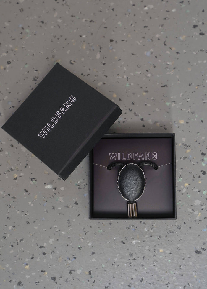 A black Wildfang brand jewelry box, shown open with its lid leaning against it, revealing a black bolo tie with circular pendant matching dark display insert, all placed on a speckled gray surface.