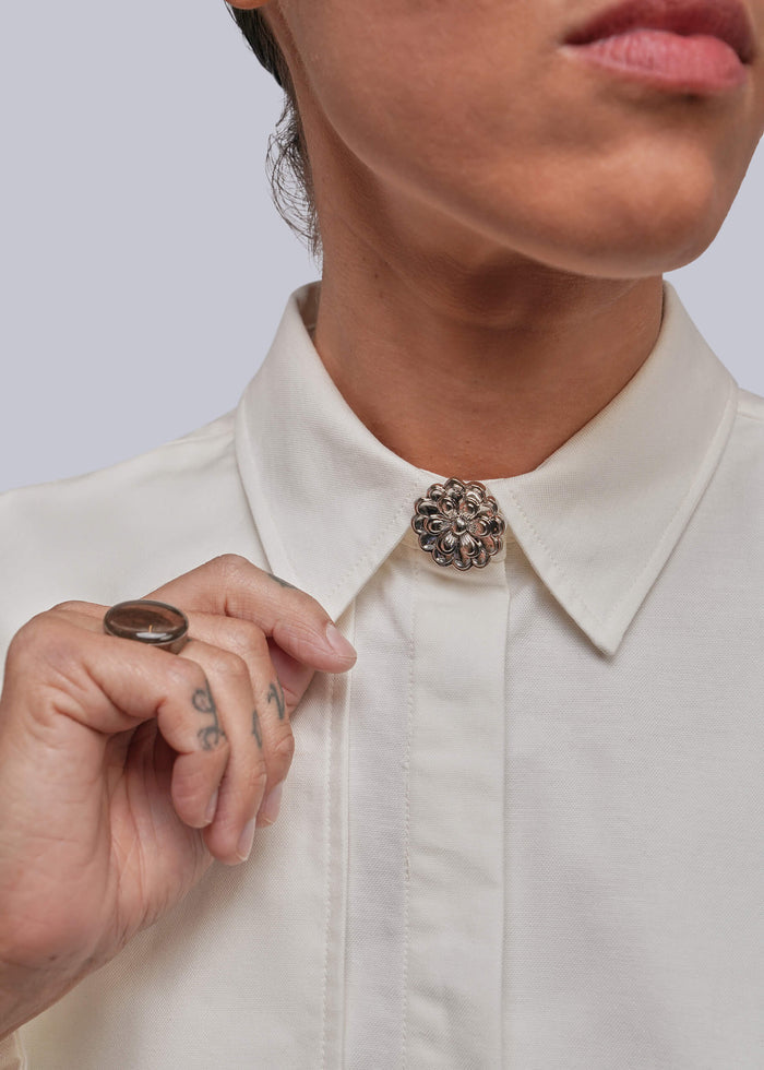 Close-up of a person wearing a white collared shirt with a decorative round silver-toned button cover at the collar, against a light gray background.