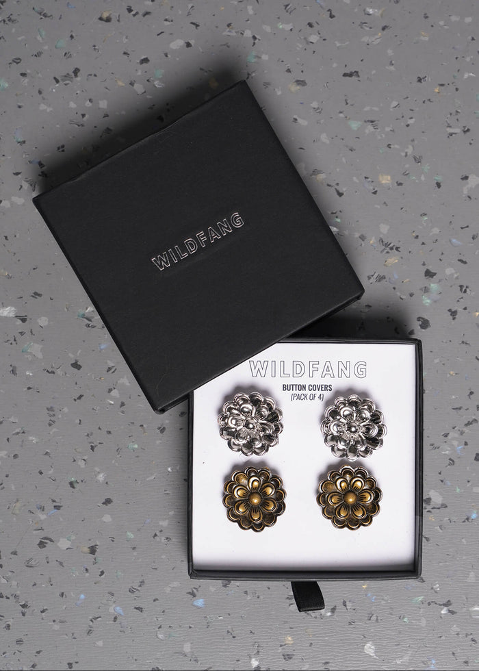 Two pairs of Wildfang button covers displayed in an open box on a speckled gray surface. One pair is silver-toned and the other is bronze-toned, with the matching black box lid placed beside it.