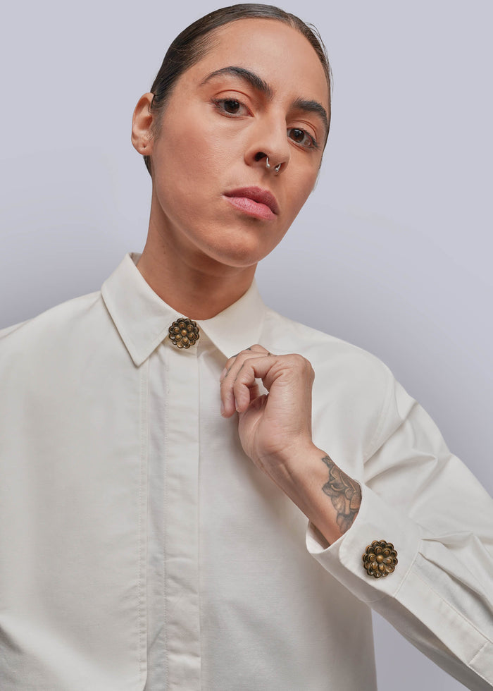 A close-up of a model wearing a white long-sleeve button-down shirt with decorative round gold-tone button covers at the collar and cuff, set against a plain light gray background.