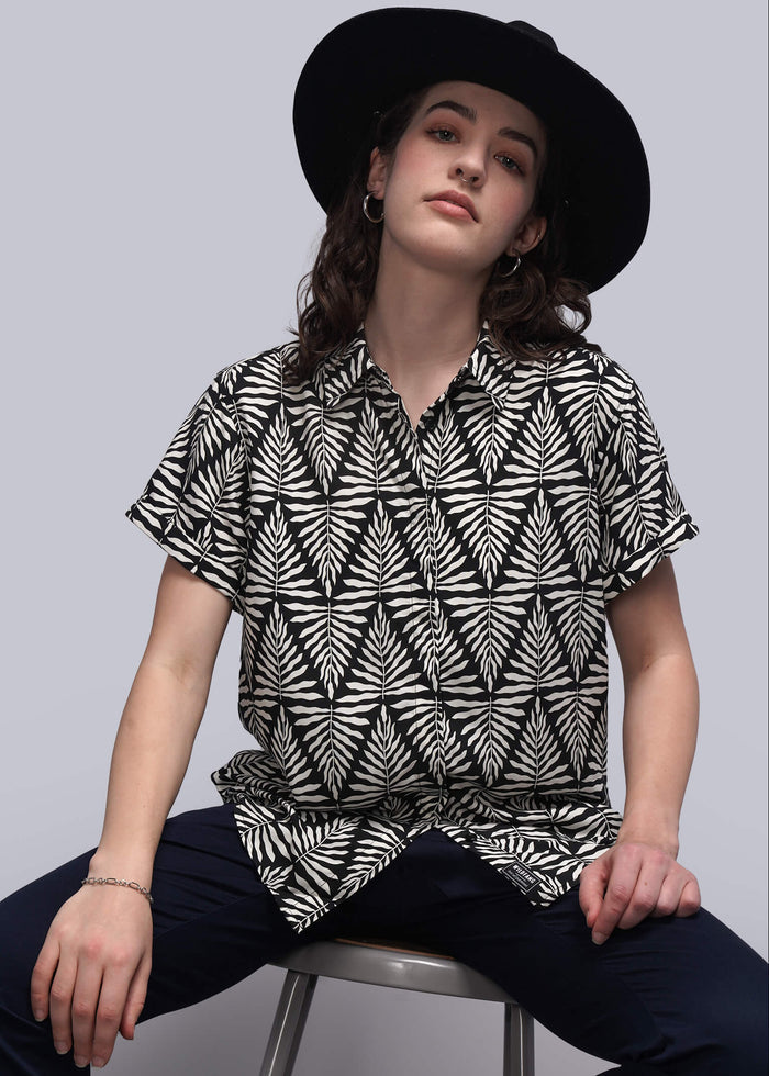 S=Person wearing a black and white patterned shirt and black hat sitting on a stool against a gray background