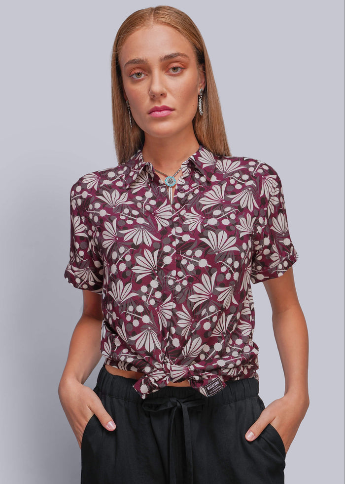 S=A woman modeling a short-sleeve button-up shirt with a burgundy and beige floral pattern, tied at the waist, paired with black drawstring pants against a solid gray background.