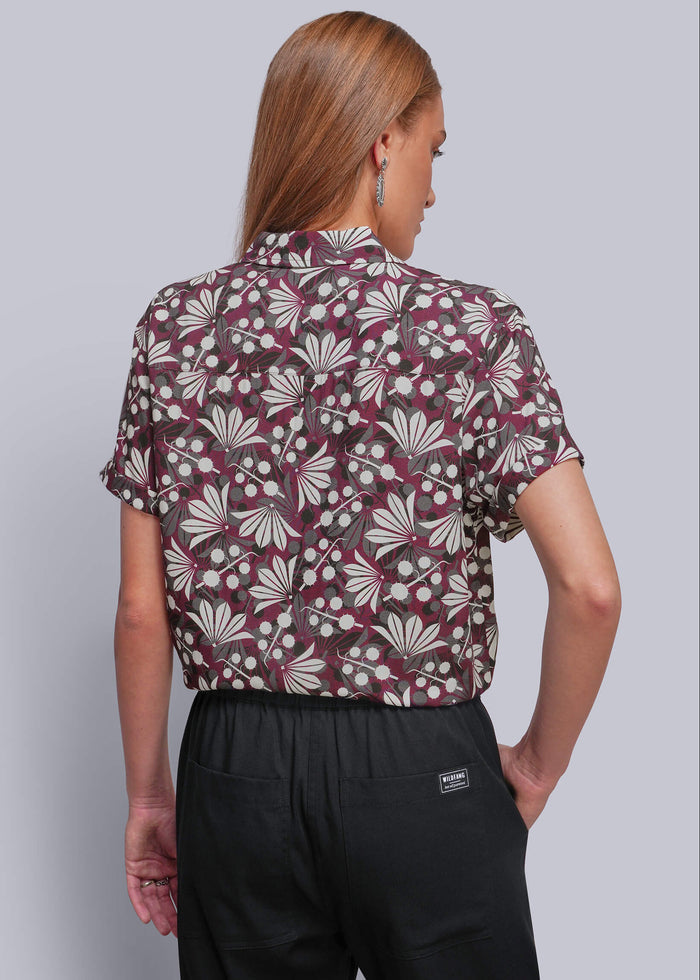S=A woman shown from the back wearing a short-sleeve button-up shirt with a burgundy, beige, and white floral pattern, paired with black trousers, against a solid gray studio background.