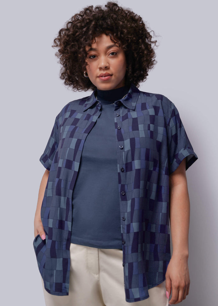 1X=Woman wearing a blue geometric patterned shirt over a plain shirt against a gray background