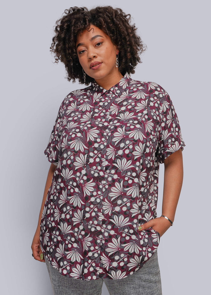 1X=A woman with curly dark hair models a short-sleeve button-up shirt featuring a burgundy, white, and gray floral pattern, paired with gray pants, against a solid light gray background.