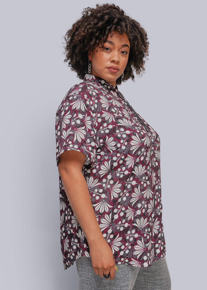 1X=A woman modeling a short-sleeve button-up shirt with a dark purple and white floral pattern, paired with gray pants, against a solid light gray background.
