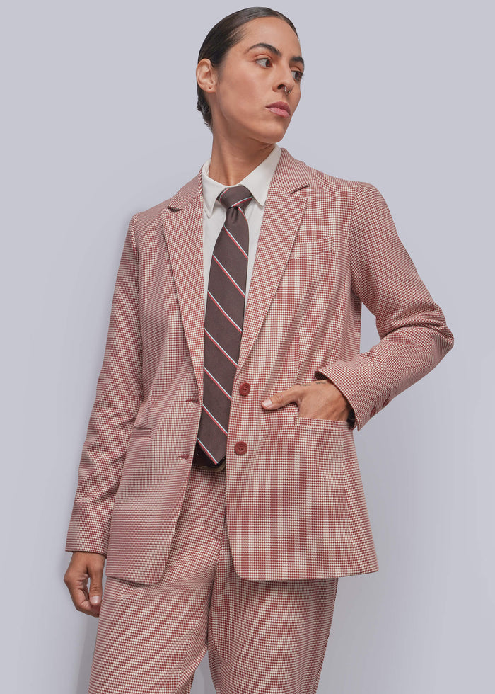 S=A model wearing a cinnamon and white micro-check suit, including a blazer and trousers, styled with a white collared shirt and a brown and pink striped tie, against a solid gray background.
