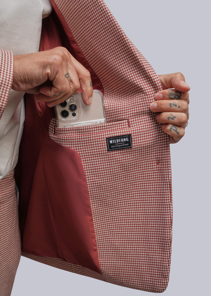 S=A close-up of a person holding open a dusty red and white micro-check blazer to show a phone pocket with a red lining. A black brand label reading WILDFANG is visible on the pocket.