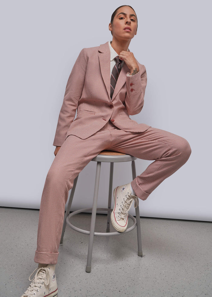 S=A model wearing a dusty red tailored suit with matching trousers, a white shirt, a striped tie, and white sneakers, sitting on a metal stool against a light gray studio background.