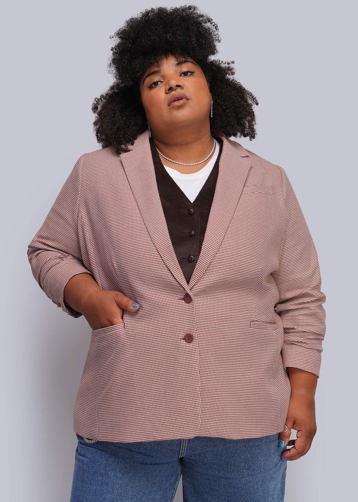 1X=A plus-size model with an afro hairstyle wearing a dusty red pink textured blazer over a dark brown vest and blue jeans, standing against a solid gray studio background.