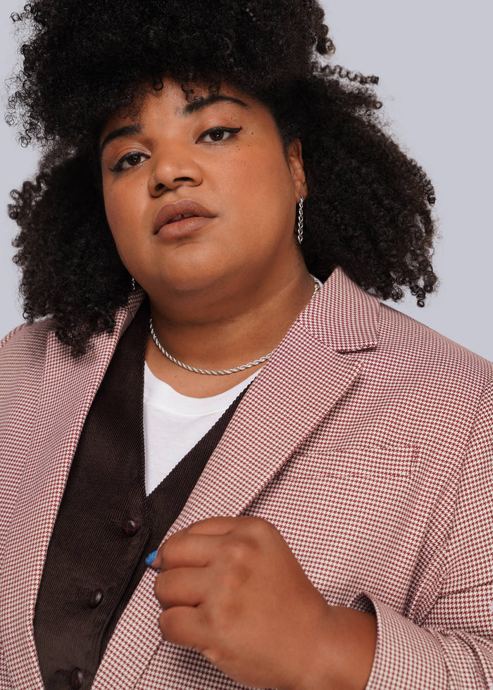 1X=A close-up portrait of a person with dark curly hair wearing a red and white micro-check blazer over a dark brown vest and white t-shirt, against a light gray background.