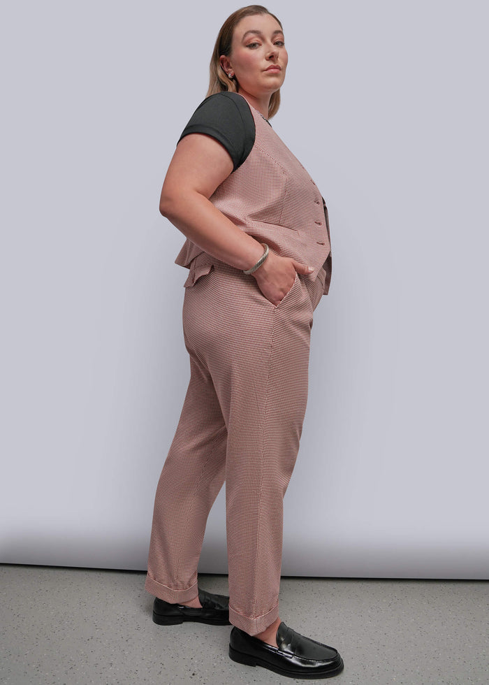1X=A woman modeling a dusty red patterned vest and matching trousers set, paired with a black short-sleeve top and black loafers, against a light gray studio background.