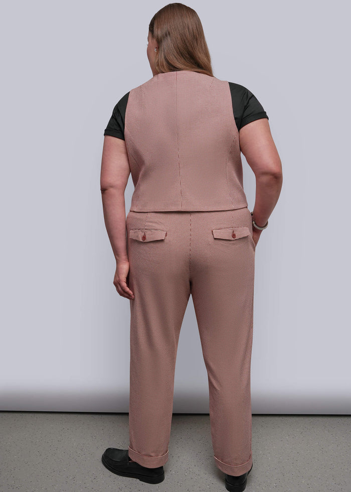 1X=A woman viewed from the back wearing a dusty red vest and matching trousers over a black short-sleeve top, standing against a plain light gray studio background.