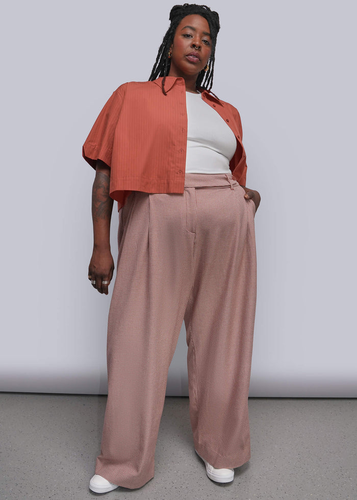 1X=A model stands against a light gray studio wall wearing a terracotta cropped button-up shirt layered over a white top and dusty red wide-leg trousers, paired with white sneakers.