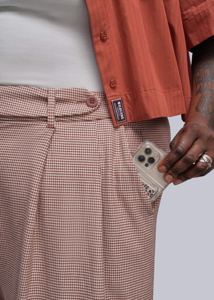 1X=Close-up of a person wearing red and beige houndstooth pleated trousers with a side pocket holding a phone, paired with a white shirt and a rust-colored short-sleeve button-up shirt. A black brand label on the shirt reads 'WILDFANG'.