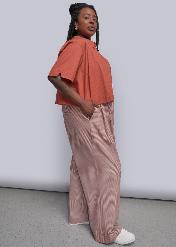 1X=A woman modeling a two-piece outfit consisting of a coral orange short-sleeve top and dusty red houndstooth wide-leg trousers, paired with white sneakers, against a plain light gray studio background.