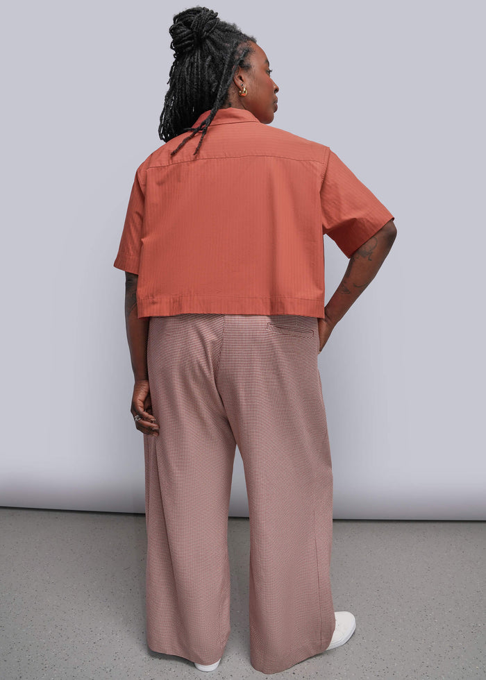 1X=A model shown from the back wearing a coral short-sleeve cropped button-up shirt and dusty red wide-leg trousers, paired with white sneakers, against a light gray studio background.